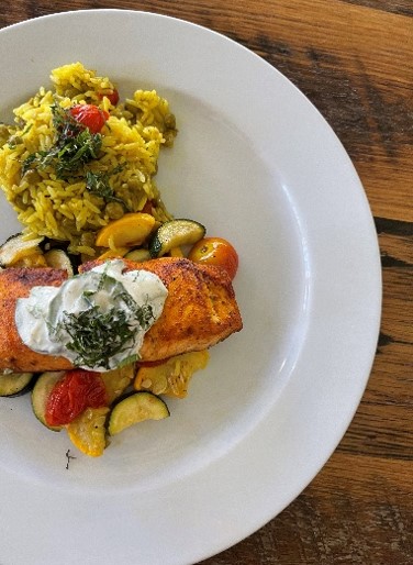 Grilled salmon with creamy sauce, cherry tomatoes, zucchini, and yellow rice served on a white plate on a wooden table.
