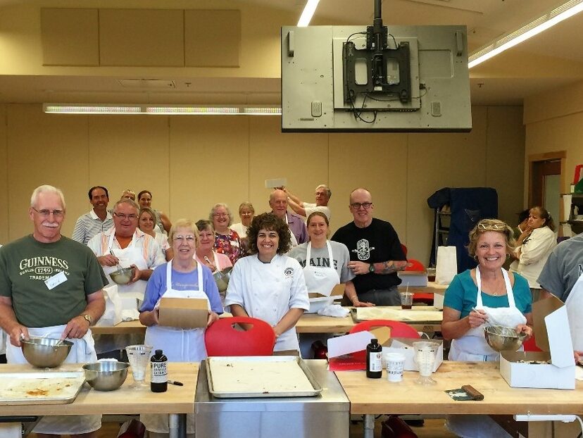 A group of adults in aprons stands smiling in a classroom kitchen, holding baking tools and supplies, with tables and baking trays in front of them.
