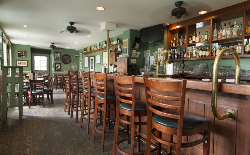 Wooden chairs line a bar in a cozy, well-lit pub with green walls, shelves of bottles, framed art, and empty tables in the background.