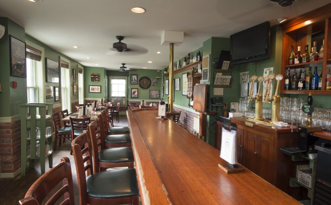 Empty bar with wooden chairs, green walls, a row of taps, shelves of bottles, and framed pictures, with natural light from windows in the background.