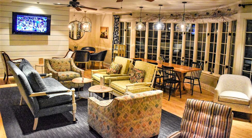 A cozy, well-lit living room with sofas, armchairs, a dining table, large windows, and a TV on the wall showing a basketball game.