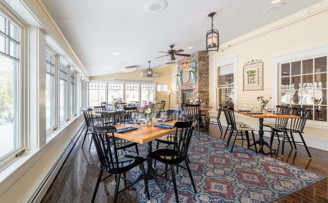 Bright restaurant with wooden tables, black chairs, large windows, floral rug, and cozy decor including wall art and a stone fireplace.