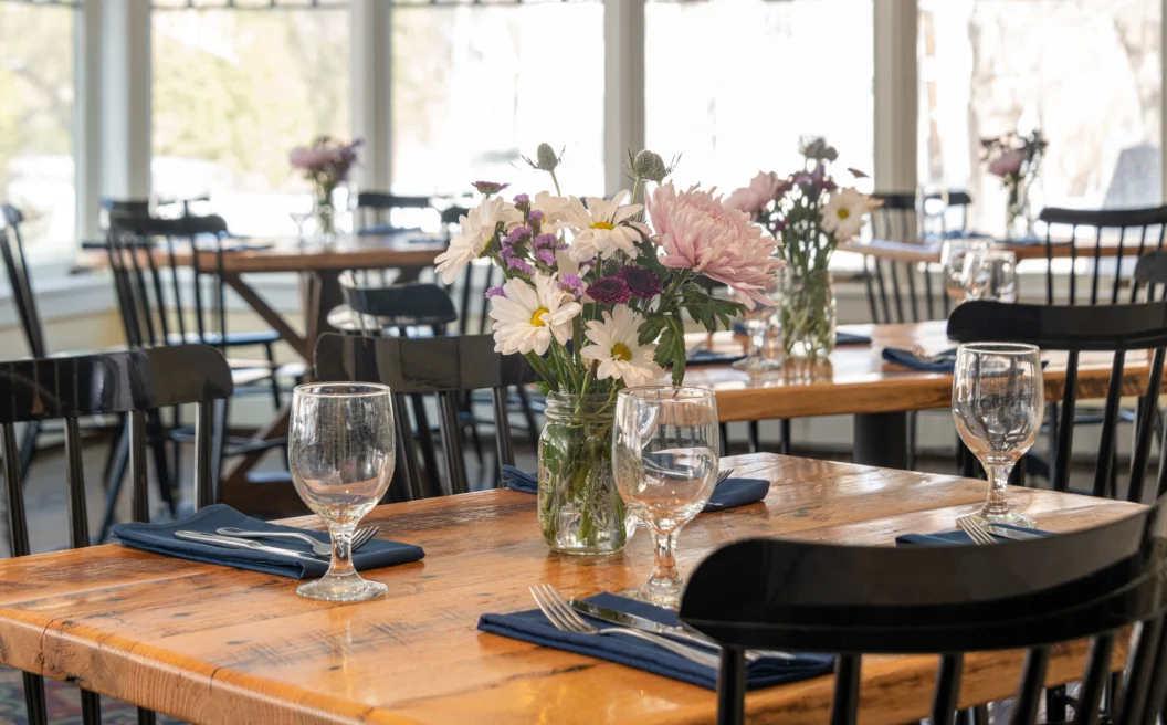 Sunlit restaurant with wooden tables, black chairs, glassware, navy napkins, silverware, and flower centerpieces in glass jars.