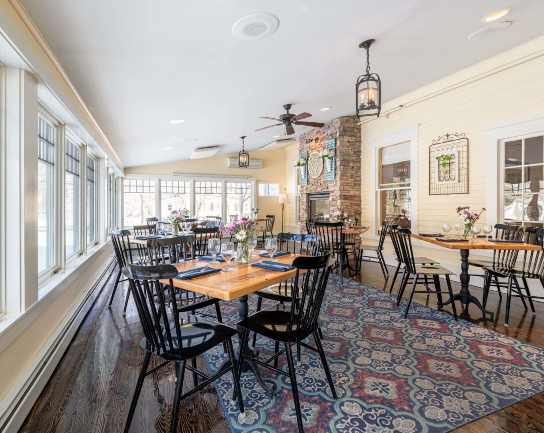 Bright restaurant with wooden tables, black chairs, large windows, floral rug, and cozy decor including wall art and a stone fireplace.