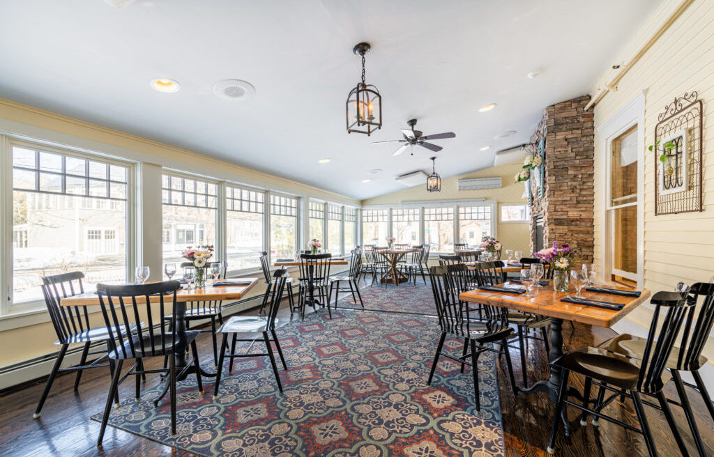 Bright, airy restaurant with black chairs, wooden tables, large windows, floral centerpieces, ceiling fans, and a patterned rug on the floor.