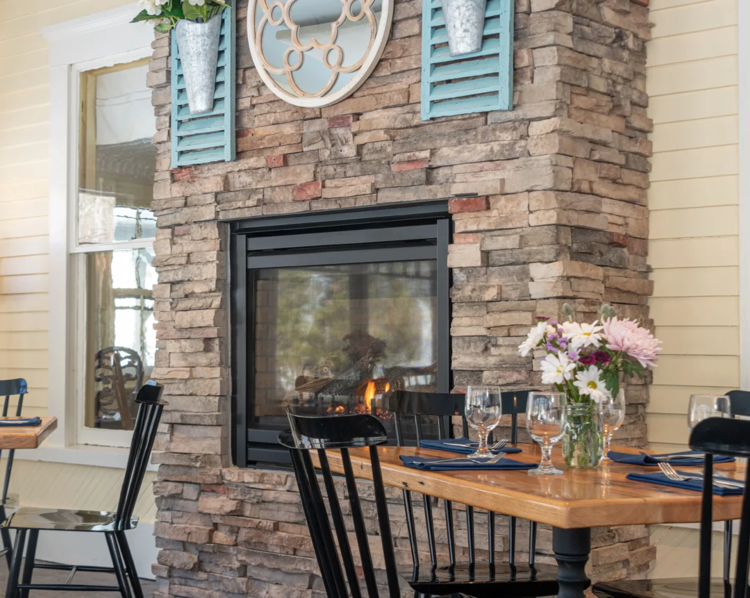 Cozy dining area with a stone fireplace, wooden chairs, and a round table set for a meal. Flowers and wall decor add a welcoming touch.