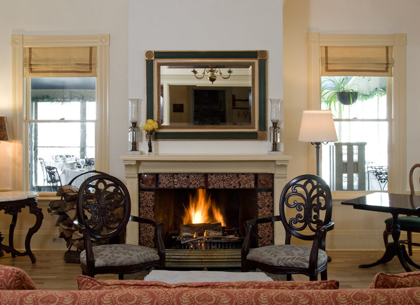 Elegant living room with a lit fireplace, two ornate chairs, a mirror above the mantel, and windows on either side with beige shades.