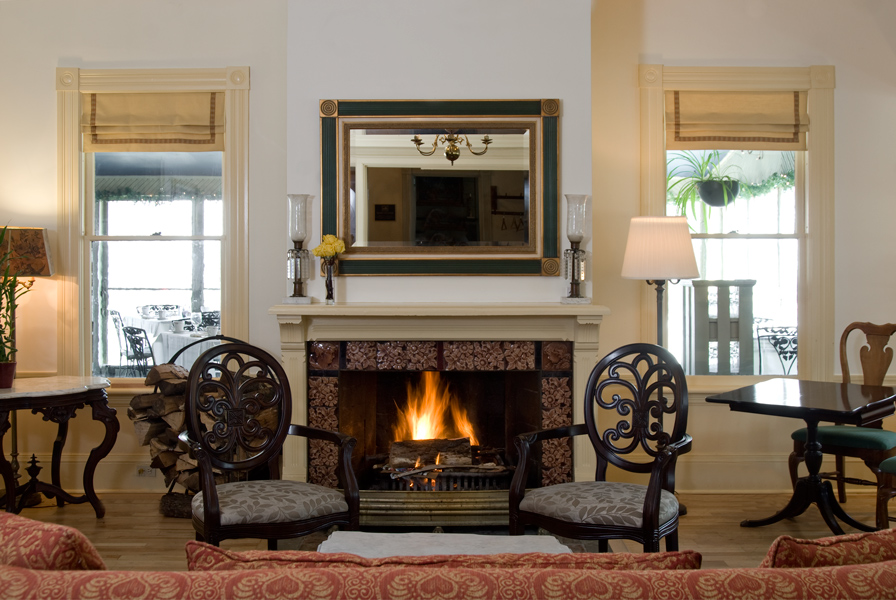 Elegant living room with a lit fireplace, two ornate chairs, a mirror above the mantel, and windows on either side with beige shades.