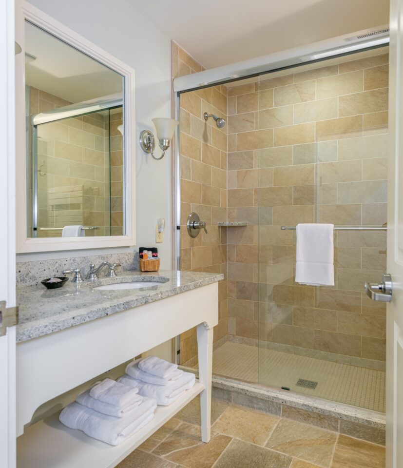 Modern bathroom with a glass shower, tan tile walls, white vanity, large mirror, and neatly folded white towels on a shelf below the sink.