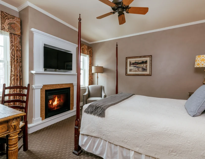 Cozy bedroom with a four-poster bed, fireplace, TV, wooden desk, armchair, and windows letting in natural light.