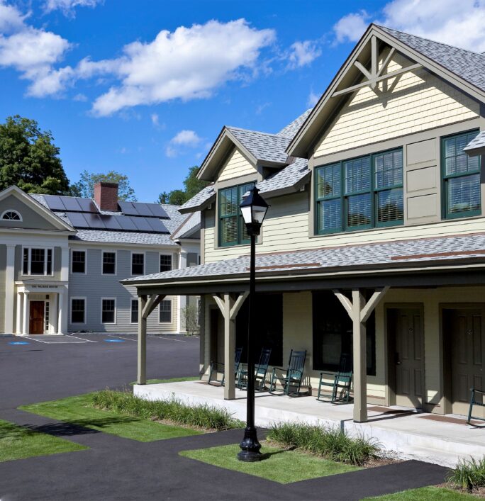 Two modern, light-colored buildings with porches and rocking chairs, under a blue sky with clouds, surrounded by green trees and pavement.
