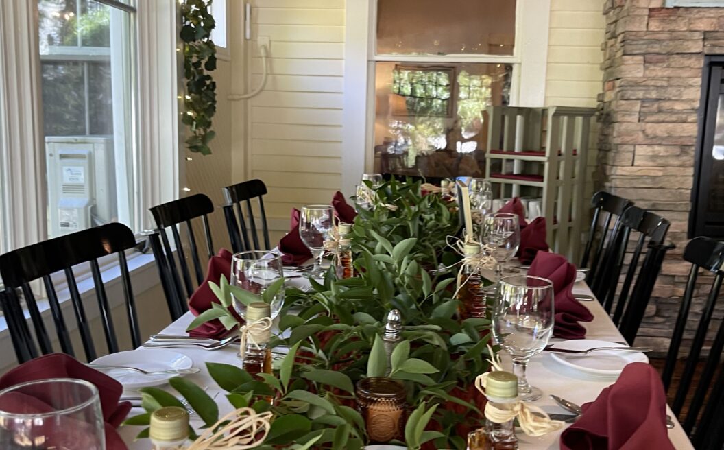 A long dining table set with wine glasses, burgundy napkins, greenery, and maple leaf favors, in a cozy, sunlit room with stone and wood walls.