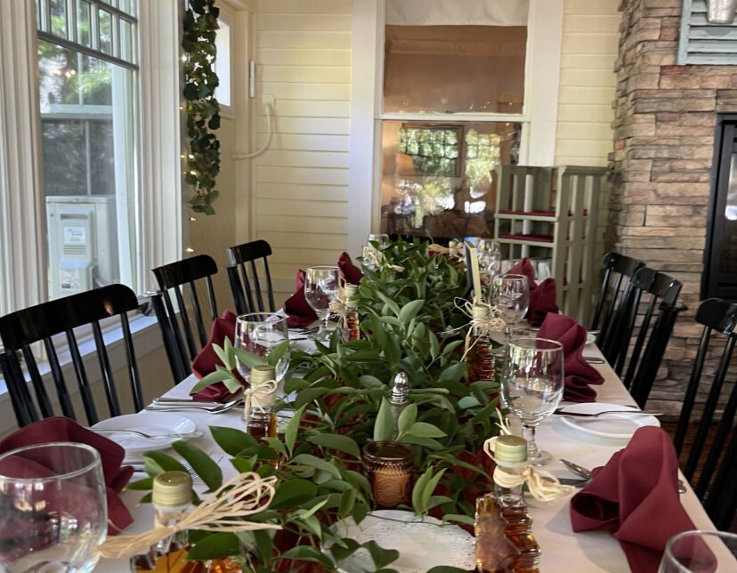 A long dining table set with wine glasses, burgundy napkins, greenery, and maple leaf favors, in a cozy, sunlit room with stone and wood walls.