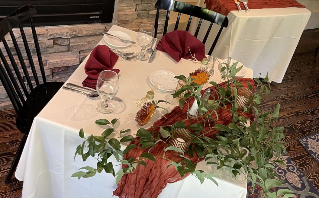 Square table set for two with burgundy napkins, greenery centerpiece, and rust-colored runner, next to a fireplace and window, on a patterned rug.