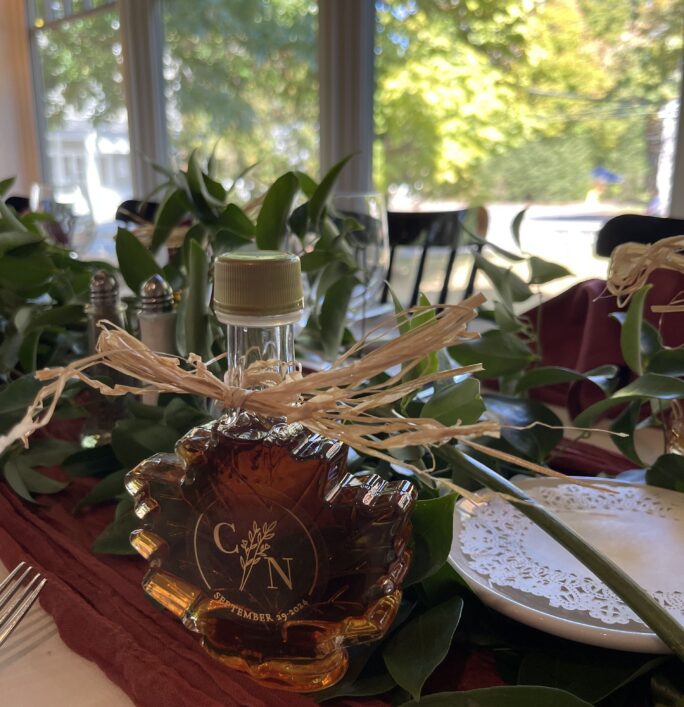 A maple leaf-shaped maple syrup bottle with a leaf design sits on a decorated table, surrounded by greenery and natural light from large windows.