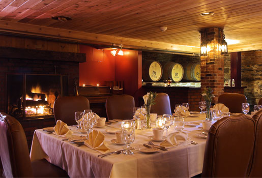 Elegant dining room with a lit fireplace, set table, wine barrels, and warm lighting creating a cozy, inviting atmosphere.