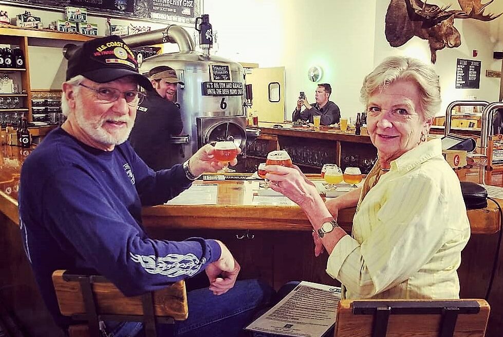 An older man and woman sit at a bar, smiling and holding drinks, with other patrons and beer taps visible in the background.