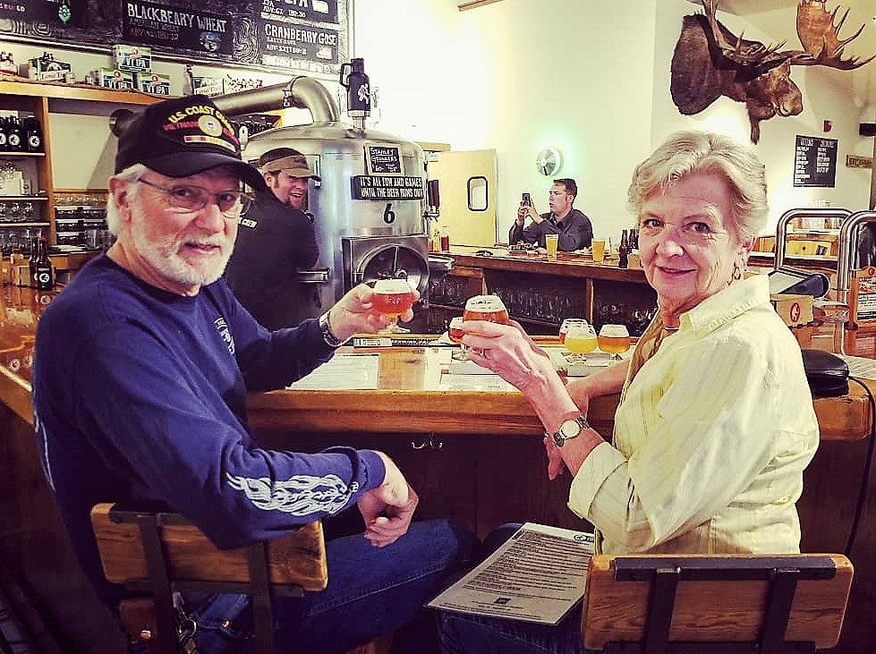 An older man and woman sit at a bar, smiling and holding drinks, with other patrons and beer taps visible in the background.