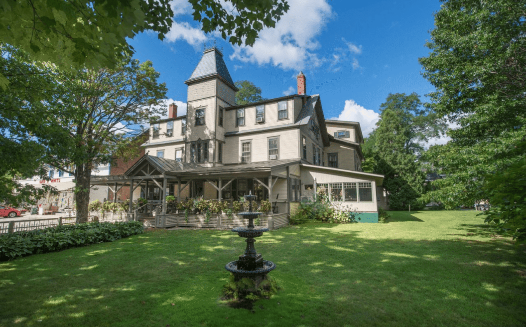 Three-story Victorian house with a tower, large porch, lawn, trees, and a black tiered fountain in front under a partly cloudy sky.