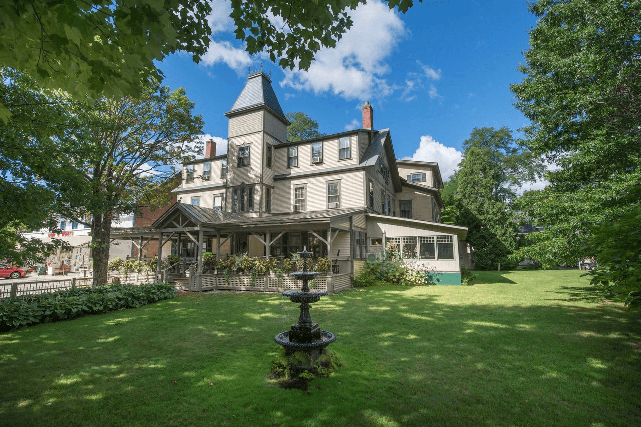 Three-story Victorian house with a tower, large porch, lawn, trees, and a black tiered fountain in front under a partly cloudy sky.