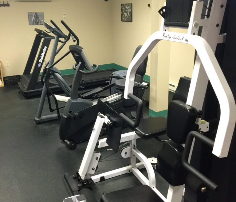A small gym with a weight machine, an exercise bike, and two treadmills on a black floor in a well-lit room.