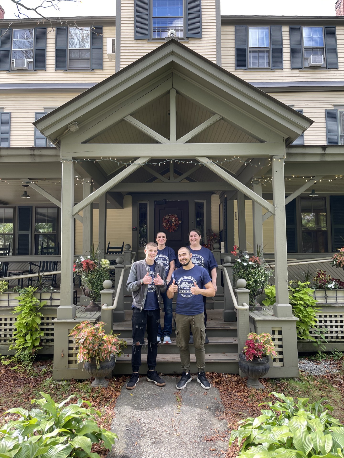 Four people stand smiling and giving thumbs up on the steps of a green house with plants and flowers, under a covered porch.
