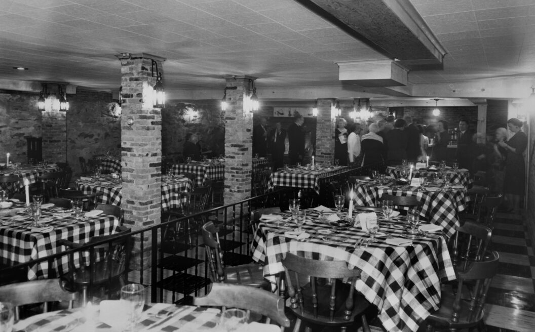 Black-and-white photo of a restaurant with checkered tablecloths, wooden chairs, brick pillars, and people standing near the back.