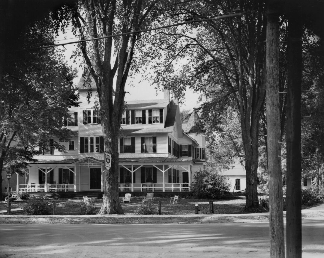 A large, white, three-story building with a porch sits behind tall trees and an empty lawn with chairs, viewed from across a road.