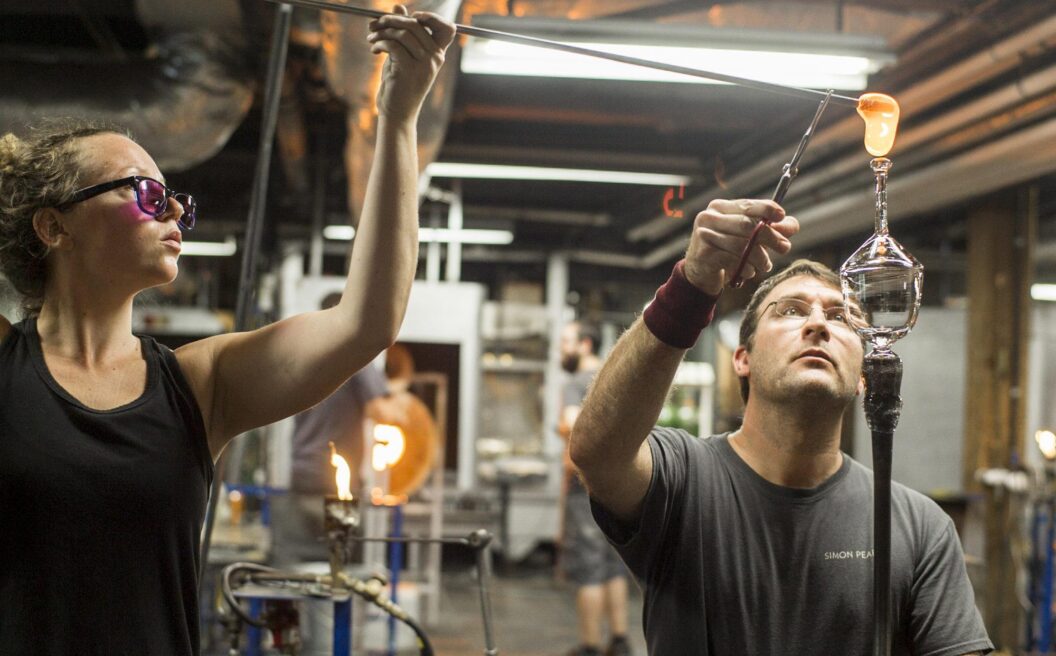 Two people work together in a glassblowing studio, carefully shaping molten glass on a rod using specialized tools, with furnaces visible in the background.