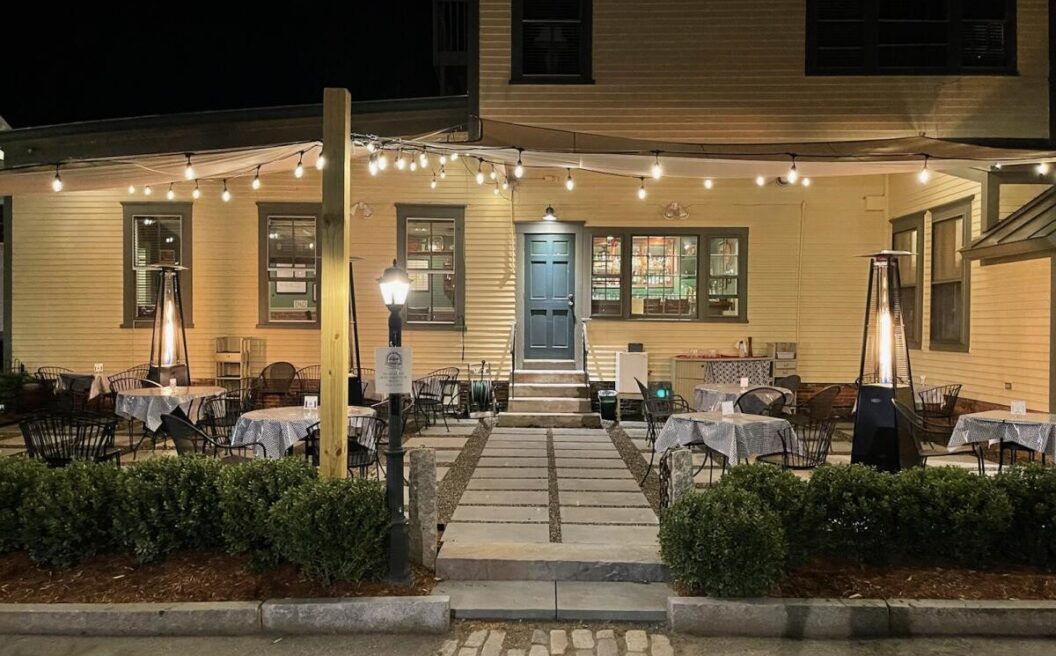 Outdoor restaurant patio at night with string lights, empty tables and chairs, heaters, and a well-lit entrance to a yellow building.