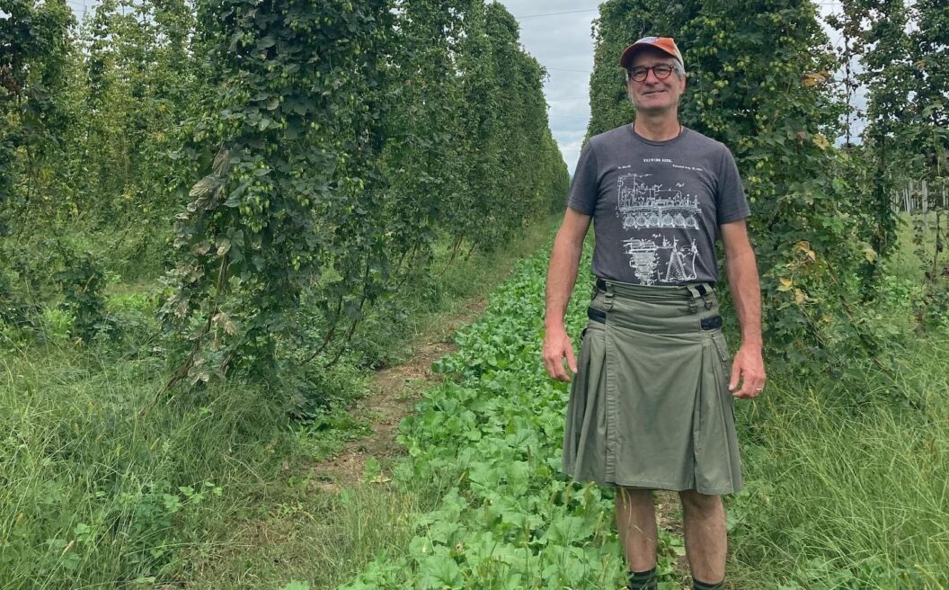 A person in a t-shirt, skirt, and boots stands smiling in a green field between tall rows of leafy plants on a cloudy day.