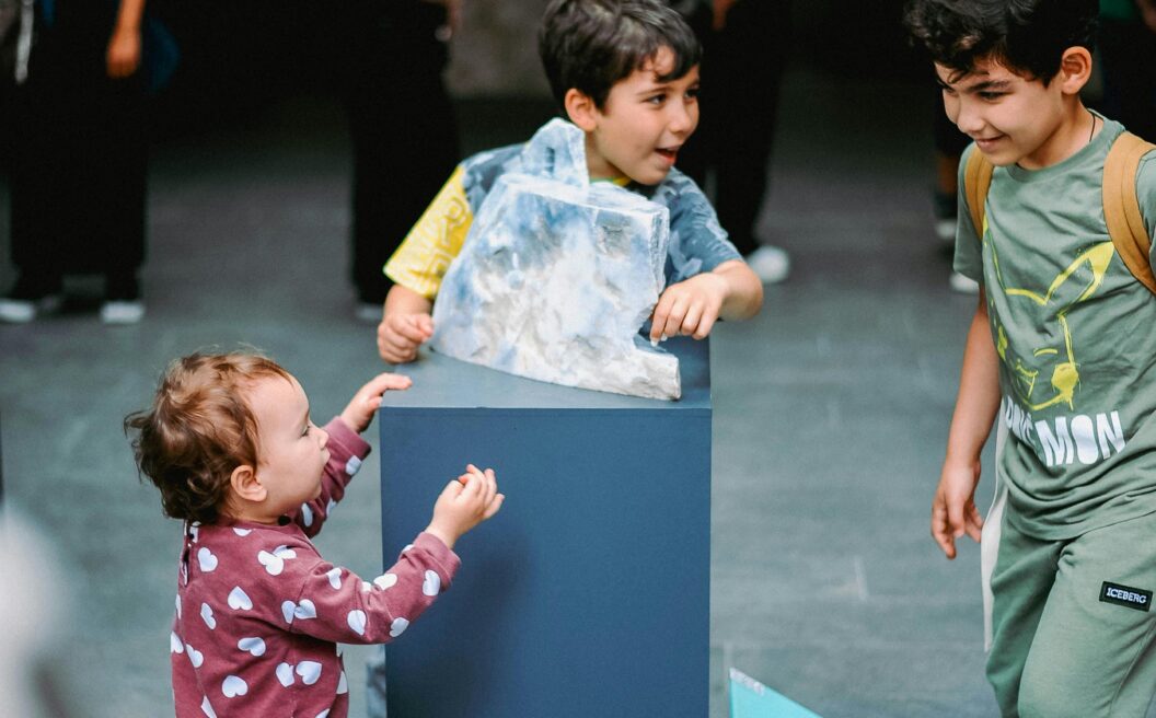 Three children interact with an exhibit in a public indoor space, while several adults stand and walk in the background.