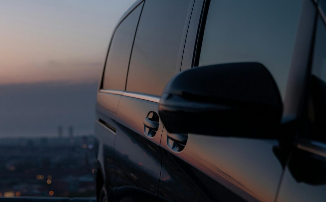 Close-up of a black van parked on a hill at sunset, city lights and sky reflecting on its windows.