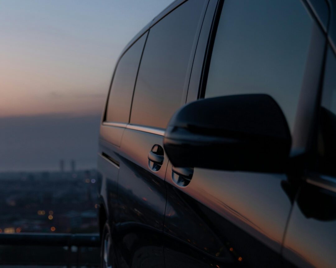 Close-up of a black van parked on a hill at sunset, city lights and sky reflecting on its windows.