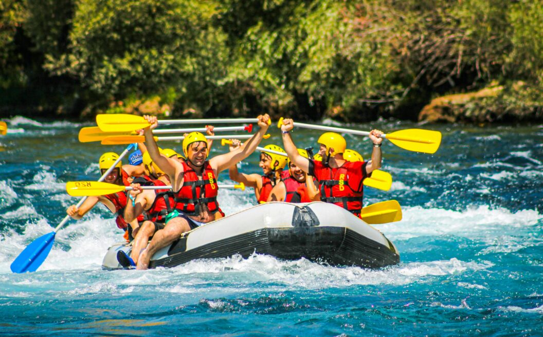A group of people wearing helmets and life jackets cheer while rafting down a river with yellow paddles, surrounded by splashing water.