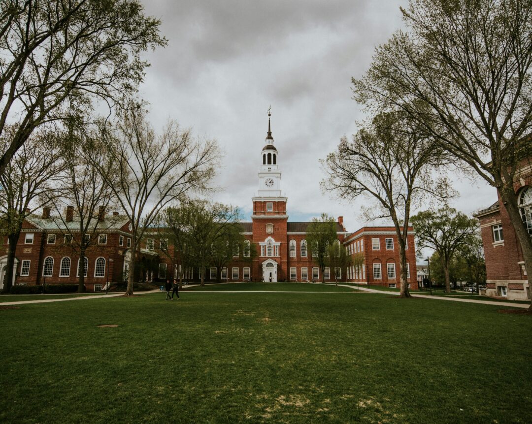 A large brick academic building with a central clock tower stands behind a green lawn, framed by trees and cloudy skies.