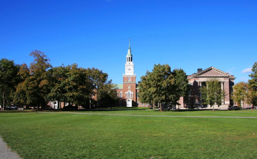 A grassy quad with trees and historic brick buildings under a clear blue sky, featuring a tall clock tower in the center.