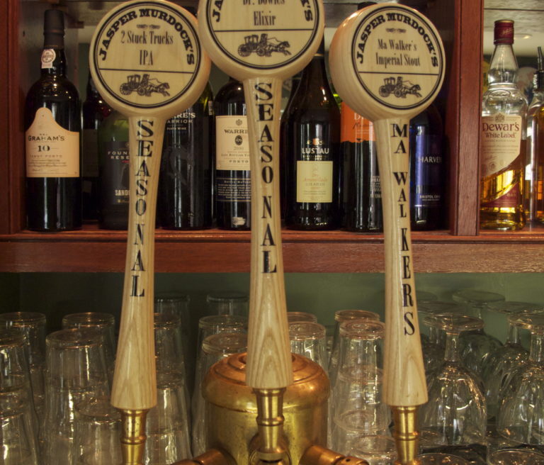 Three beer taps labeled Jasper Murdocks, surrounded by liquor bottles and glassware on shelves in a bar setting.