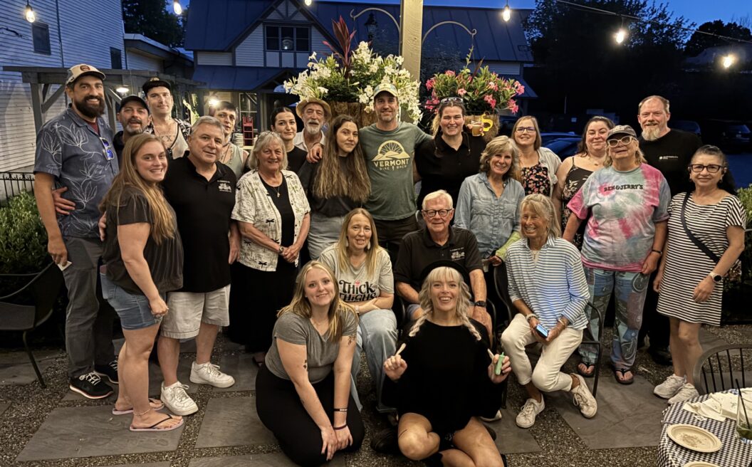A group of people of various ages pose and smile together outdoors at night under string lights, with flowers and trees in the background.