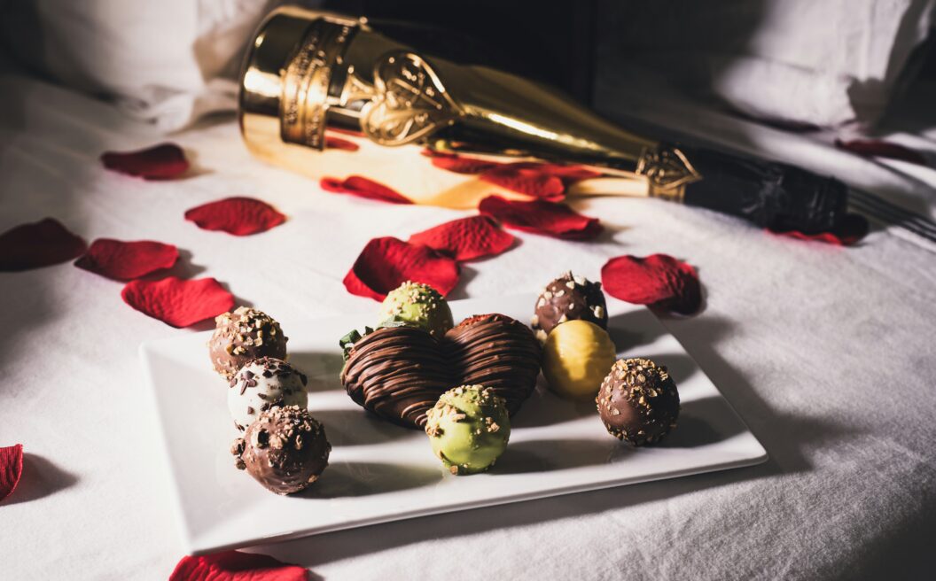 Chocolate truffles and a heart-shaped treat on a white plate, with rose petals and a gold champagne bottle on a white bed.